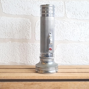 Könnte beinhalten: Eine silberne Vintage-Taschenlampe mit geripptem Gehäuse und rotem Knopf. Die Taschenlampe hat einen breiten Sockel und einen Schalter an der Seite. Sie ist aus Metall und hat ein klassisches Design.