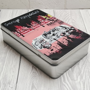 Pode incluir: Uma lata de metal prateada com um design gráfico preto e rosa. O design apresenta um carro Porsche 911 branco e o texto "Garage Keepsakes", "Owners Works", "Top Me" e "Porsche 911".