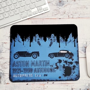 May include: A blue and black mousepad with a vintage car design. The mousepad features two black silhouettes of Aston Martin cars and the text "ASTON MARTIN 1921-1958 AUTOBOOK AUTOPRESS LTD 446".