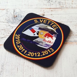 May include: A blue and yellow round coaster with a black and white image of a racing helmet. The helmet has the words "Red Bull" and "Arai" on it. The text around the helmet reads "S. Vettel 2010, 2011, 2012, 2013".