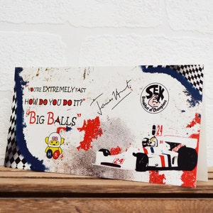 May include: A white card with a blue and red checkered border. The card features a cartoon illustration of a yellow bear wearing a racing helmet and a Union Jack flag. The text "YOU'RE EXTREMELY FAST HOW DO YOU DO IT?"BIG BALLS" is printed on the card. The card is signed by James Hunt.