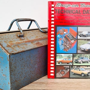 Op de afbeelding: Een blauwe metalen gereedschapskist met een roestige patina en een rode Snap-on Tools technische datamanual uit 1983. De handleiding heeft een spiraalbinding en bevat foto's van auto's en gereedschap.