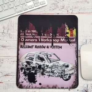 Puede incluir: Alfombrilla de ratón rectangular con un esquema de coche antiguo y el texto "Owners Workshop Manual RELIANT ROBIN & KITTEN". El diseño incluye una ilustración de coche y un fondo desgastado de estilo grunge con salpicaduras de pintura negra. Un ratón y un teclado blancos están en el fondo.