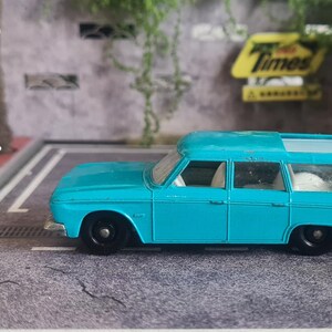 May include: A teal blue vintage station wagon toy car with black wheels. A white figurine of a person aiming a gun stands near a Matchbox box. A white dog figurine stands near the car.