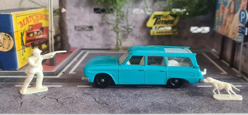 May include: A teal blue vintage station wagon toy car with black wheels. A white figurine of a person aiming a gun stands near a Matchbox box. A white dog figurine stands near the car.