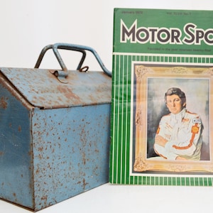 May include: A blue metal toolbox with a rusty patina and a handle. The toolbox is next to a vintage magazine called "Motor Sport" with a green cover and a photo of a race car driver on the front.