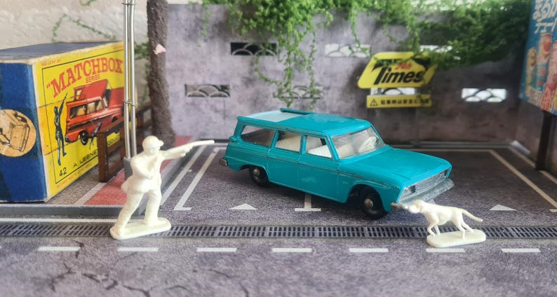 May include: A turquoise toy estate car with white interior, a white dog, and a white figure. A Matchbox box is on the left. The scene is set on a grey surface with white lines, with a Times sign in the background.