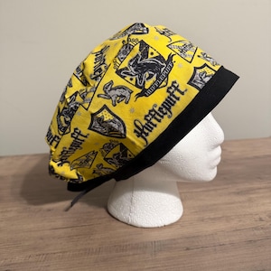 Könnte beinhalten: Eine gelbe OP-Haube mit schwarzem Rand. Die Haube ist mit schwarzen Hufflepuff-Wappen und Text bedruckt. Die Haube wird hinten mit schwarzen Bändern gebunden. Die Haube wird auf einem weißen Schaumstoffkopf präsentiert.