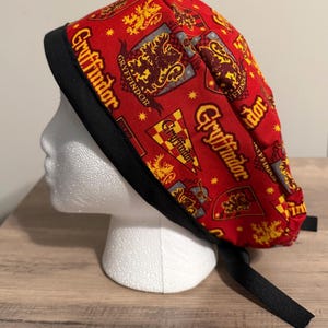 May include: A red and gold patterned scrub cap featuring the word "Gryffindor" and a crest design. The cap has a black band and ties. The fabric is printed with the Gryffindor house crest and other related imagery.