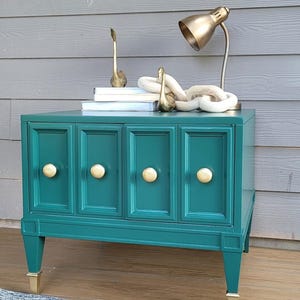 May include: Teal-colored cabinet with four doors and gold knobs. The cabinet has gold-tipped legs and sits on a wooden surface. Decorative items on top include a gold lamp, swan figurines, and books. A blue and white rug is in front.