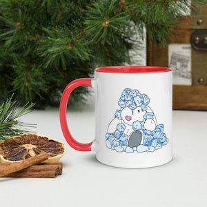 Könnte beinhalten: Weiße Keramiktasse mit rotem Rand. Die Tasse zeigt eine Cartoon-Illustration eines weißen Bären, der einen blauen Hut trägt und in einem Haufen blauer Schneebälle sitzt.