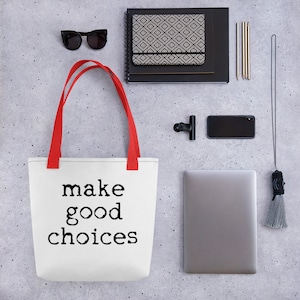 Puede incluir: Bolsa de tela blanca con asas rojas y el texto "make good choices" impreso en negro. La bolsa está sobre un fondo de hormigón gris con otros artículos, como un portátil, una libreta, gafas de sol y un teléfono.