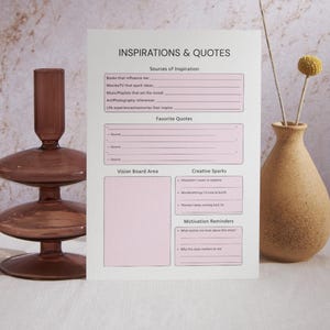 May include: A white paper titled "INSPIRATIONS & QUOTES" with pink boxes for notes. A brown glass candle holder and a beige vase with dried flowers are on either side of the paper.