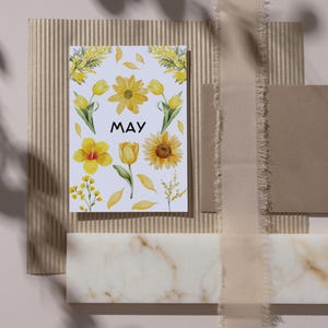 May include: A white card with the word "MAY" in black text and yellow watercolor flowers, including tulips, sunflowers, and mimosa. The card is on a brown corrugated cardboard surface.