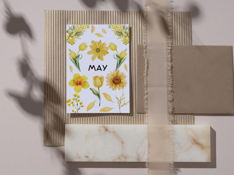 May include: A white card with the word "MAY" in black text and yellow watercolor flowers, including tulips, sunflowers, and mimosa. The card is on a brown corrugated cardboard surface.