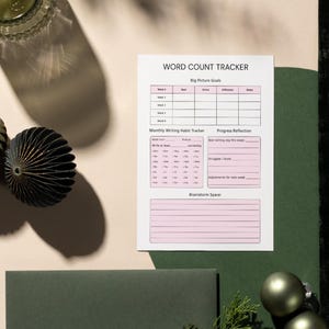 May include: A Word Count Tracker sheet with pink accents lies on a light beige surface. The sheet includes sections for weekly goals, a monthly writing habit tracker, and a space for brainstorming. Decorative paper ornaments and a glass are also visible.