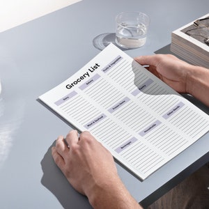 May include: A person holding a printed "Grocery List" on a light gray surface. The list is divided into sections like "Dairy," "Fruits & Vegetables," and "Frozen Items." A glass of water and a decorative glass container are also visible.