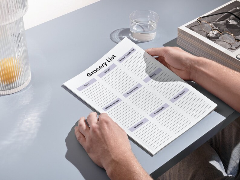 May include: A person holding a printed "Grocery List" on a light gray surface. The list is divided into sections like "Dairy," "Fruits & Vegetables," and "Frozen Items." A glass of water and a decorative glass container are also visible.