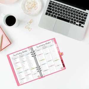 May include: A pink travel packing checklist with a silver binder ring. The checklist is open to two pages, each with a list of items to pack for a trip. The checklist is on a white surface with a laptop, a cup of coffee, and other office supplies.