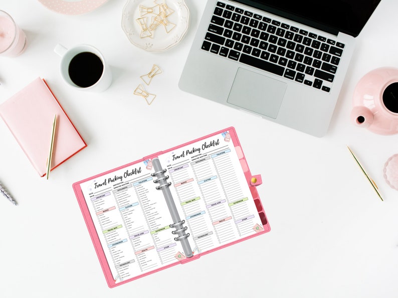 May include: A pink travel packing checklist with a silver binder ring. The checklist is open to two pages, each with a list of items to pack for a trip. The checklist is on a white surface with a laptop, a cup of coffee, and other office supplies.