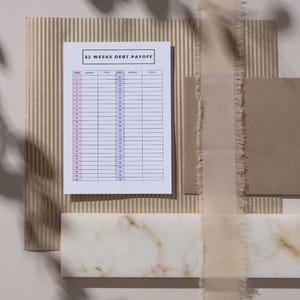 May include: A flat lay featuring a 52 Weeks Debt Payoff chart, a beige envelope, and a decorative marble piece. The chart is white with pink and black text. A beige ribbon and corrugated paper add texture to the composition.