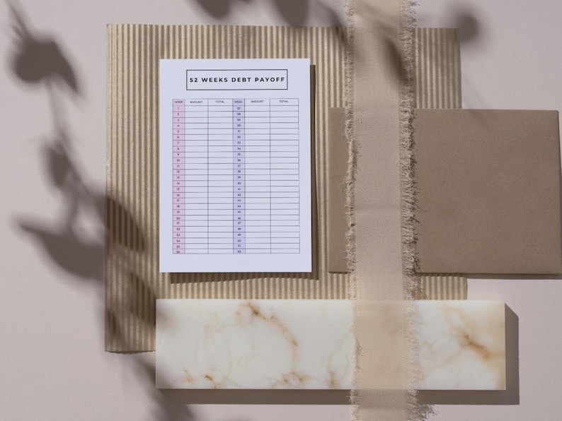 May include: A flat lay featuring a 52 Weeks Debt Payoff chart, a beige envelope, and a decorative marble piece. The chart is white with pink and black text. A beige ribbon and corrugated paper add texture to the composition.