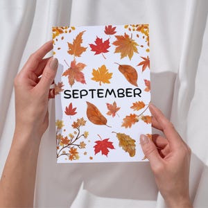 May include: A white card with the word "SEPTEMBER" printed in black. The card is decorated with a pattern of orange, yellow, and red autumn leaves.