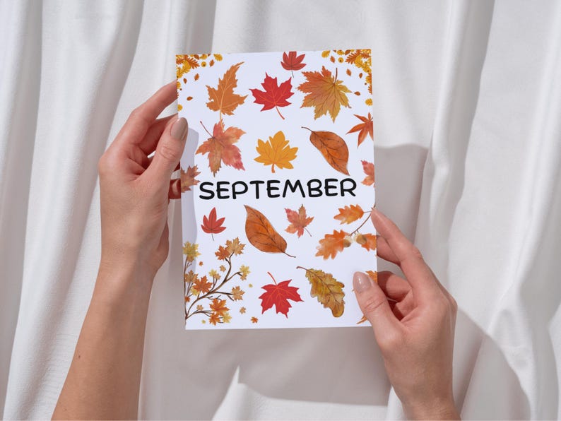 May include: A white card with the word "SEPTEMBER" printed in black. The card is decorated with a pattern of orange, yellow, and red autumn leaves.