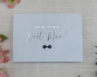 May include: A light blue card with the text "TOM. WILL YOU BE MY Best Man?" in silver foil. A small black bow tie graphic is below the text. The card is on a neutral background.