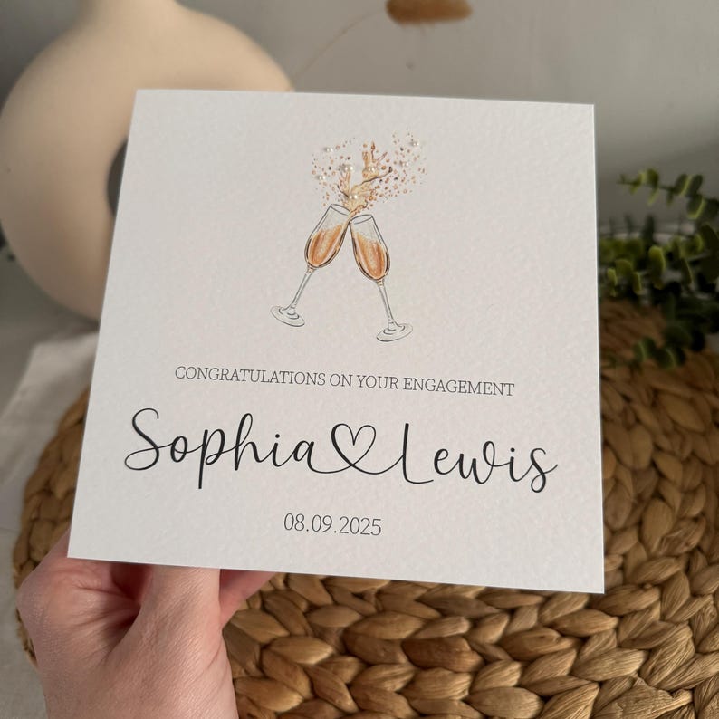 May include: A white engagement card with the text "Congratulations on your engagement" and the names "Sophia & Lewis" in elegant script. Two champagne glasses are clinking, with gold confetti. The date "08.09.2025" is also printed on the card.