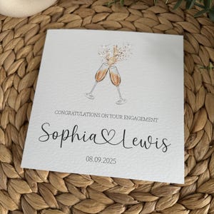 May include: A white engagement card with the text "Congratulations on Your Engagement" and the names "Sophia & Lewis" in elegant script. Two champagne glasses are clinking at the top, with the date "08.09.2025" below the names.