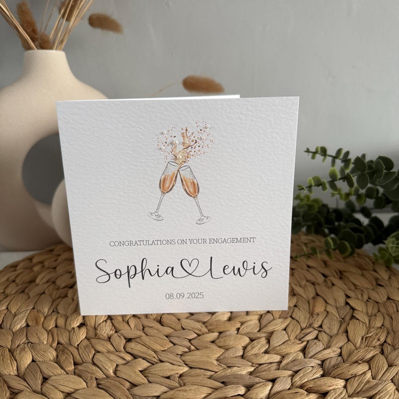 May include: A white engagement card with the text "Congratulations on your engagement" and the names "Sophia & Lewis" below an illustration of two champagne glasses clinking. The date "08.09.2025" is printed below the names.