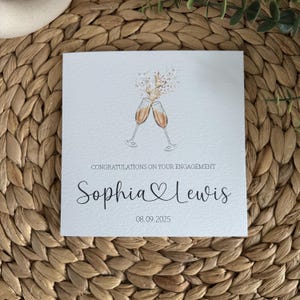 May include: Square engagement card with two champagne glasses clinking, with the text "Congratulations on your engagement". The names "Sophia & Lewis" are written below, along with the date "08.09.2025".