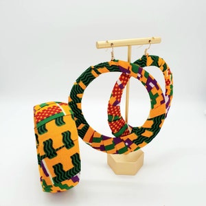 May include: A pair of large hoop earrings and a matching bracelet made of colourful fabric. The earrings are yellow, green, purple, and red with a geometric pattern. The bracelet is the same pattern and colours.