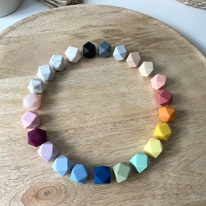 May include: A circle of 18 colorful silicone teething beads in various shades of blue, green, yellow, orange, pink, red, and gray. The beads are arranged in a circle on a wooden surface.