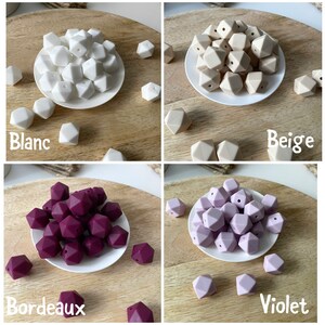 May include: Four sets of silicone teething beads in different colors: white, beige, burgundy, and lavender. Each set is displayed on a white plate on a wooden surface. The colors are labeled "Blanc", "Beige", "Bordeaux", and "Violet".