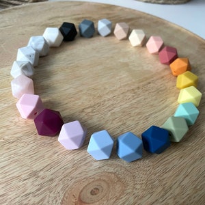 May include: A rainbow of colors in a circle of silicone teething beads. The beads are shaped like a dodecahedron and have a hole in the center. The beads are arranged in a circle on a wooden surface.