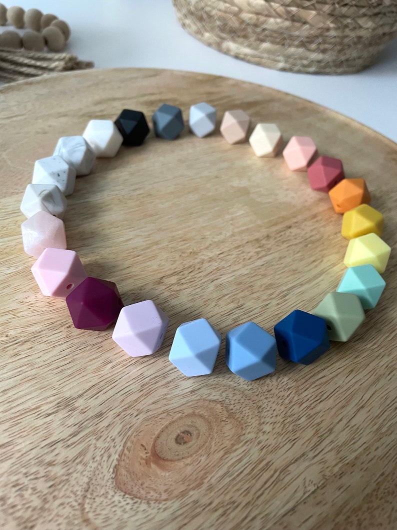 May include: A rainbow of colors in a circle of silicone teething beads. The beads are shaped like a dodecahedron and have a hole in the center. The beads are arranged in a circle on a wooden surface.