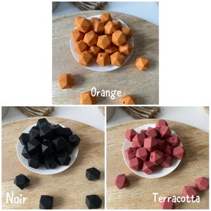 May include: Three sets of silicone teething beads in orange, black, and terracotta. Each set is arranged in a pile on a small white plate. The plates are on a wooden surface. The beads are geometric and have a faceted design.