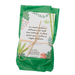 May include: A green tissue paper package tied with a brown string. The package contains a card with a message about waking up children. The card has a jungle theme with green leaves, bamboo, and pink flowers. The text on the card reads: "Le r&eacute;veille plus efficace, ce sort mes enfart. Il faut juste que arrive &agrave; r&eacute;gler Meure." The card also has the text "Les Petits Culottes La French Couche".