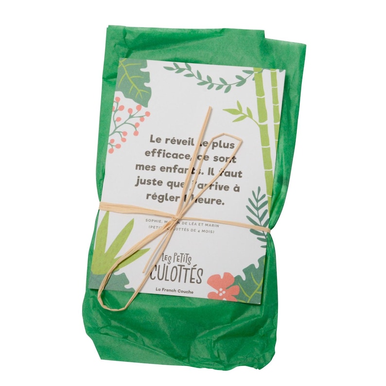 May include: A green tissue paper package tied with a brown string. The package contains a card with a message about waking up children. The card has a jungle theme with green leaves, bamboo, and pink flowers. The text on the card reads: "Le r&eacute;veille plus efficace, ce sort mes enfart. Il faut juste que arrive &agrave; r&eacute;gler Meure." The card also has the text "Les Petits Culottes La French Couche".