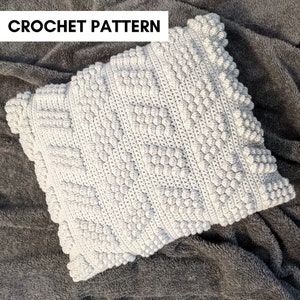 Puede incluir: Una almohada blanca de crochet con un patrón texturizado en relieve. La almohada está hecha con un hilo grueso y tiene una forma cuadrada.