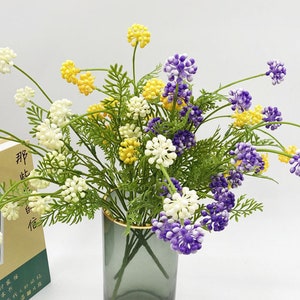 May include: A bouquet of artificial flowers in a clear glass vase. The flowers are white, yellow, and purple. The vase has a gold rim.
