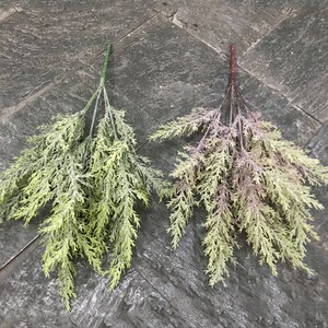 Puede incluir: Dos ramitas de vegetaci&oacute;n artificial, una verde y otra con un tono morado, sobre una superficie gris texturizada.