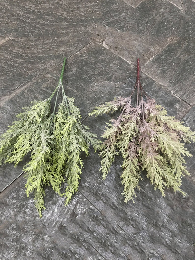 Puede incluir: Dos ramitas de vegetaci&oacute;n artificial, una verde y otra con un tono morado, sobre una superficie gris texturizada.