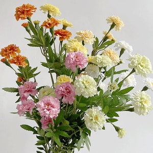 Puede incluir: Un ramo de flores artificiales en tonos naranja, amarillo, rosa y blanco. Las flores son caléndulas y tienen un aspecto realista.