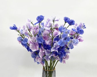 May include: A bouquet of purple and blue sweet pea flowers in a clear glass vase with a gold rim.