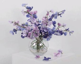 May include: A bouquet of purple and blue sweet pea flowers in a clear glass vase with a decorative wire design. The flowers are arranged in a loose, natural style.
