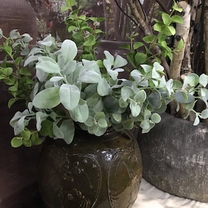 May include: A green ceramic vase with a textured surface holds a bouquet of green and white foliage. The vase is sitting on a white surface.