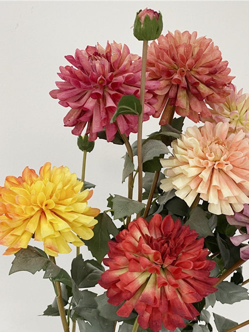 May include: A bouquet of artificial flowers with pink, yellow, and red dahlias. The flowers are arranged in a vase and have green stems and leaves.